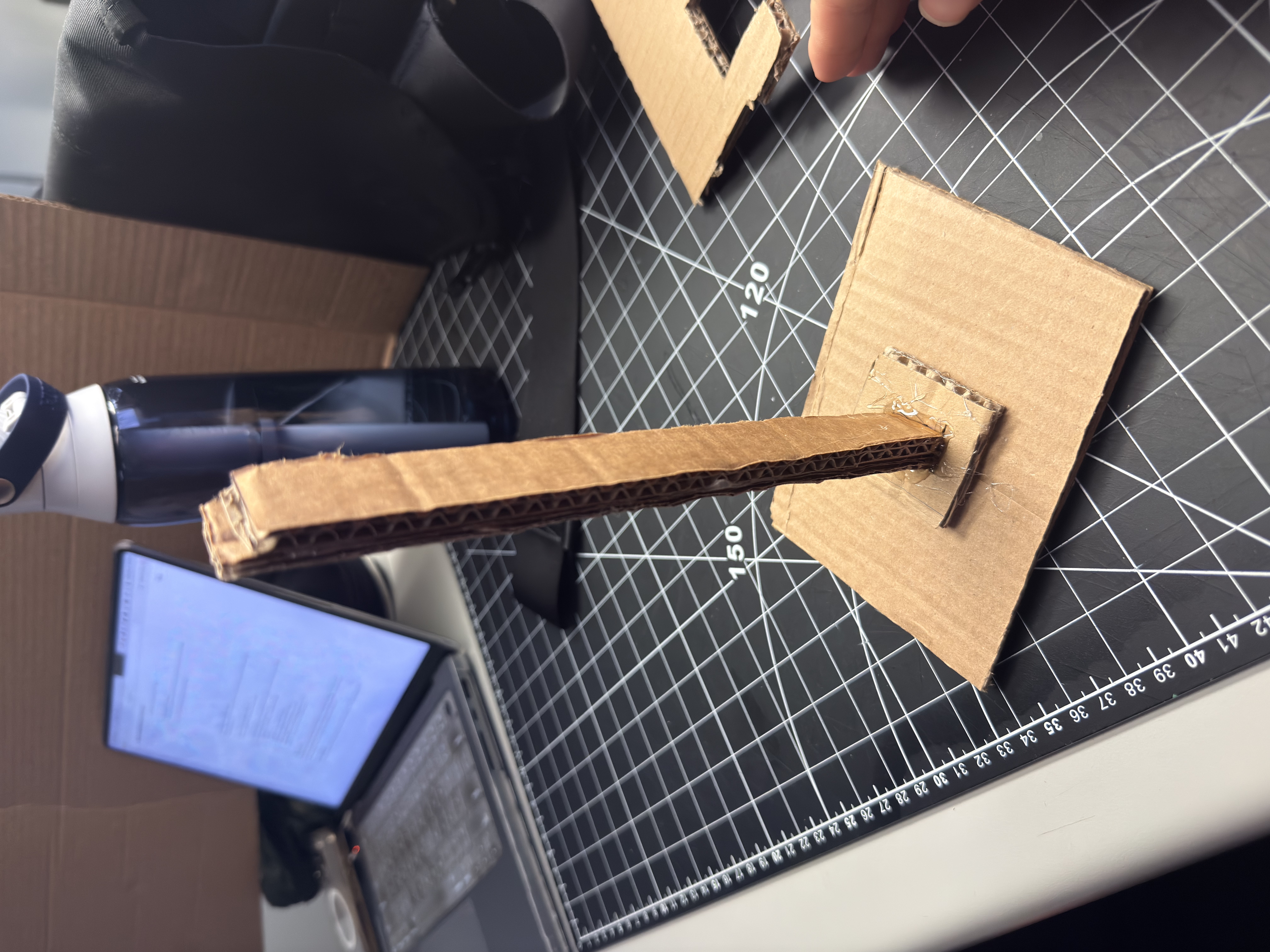 Step 1 — V1 skeleton: cardboard base + slim post to quickly test center of mass and load. Issue: wobble at the post–base joint; fix: enlarge base or add triangular bracing. Materials: corrugated cardboard, light hot-glue tacks.